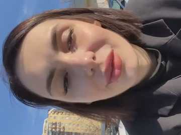 Media: Video of a young Asian woman with fair skin, straight brown hair, and light makeup, smiling warmly. She wears a black coat. The background features a clear blue sky and blurred, yellowish buildings, suggesting an urban outdoor setting.