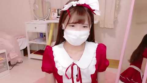 Media: Video of an Asian woman in a red maid outfit with white frills, wearing a white face mask, standing in a pink-themed room with a bed, mirror, and shelves.