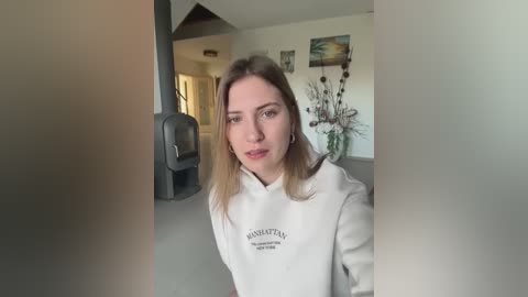 Media: Video of a blonde woman with light skin, wearing a white sweatshirt with \"HANDBAG\" text, standing in a modern, minimalist living room with beige walls, a fireplace, and a decorative vase.