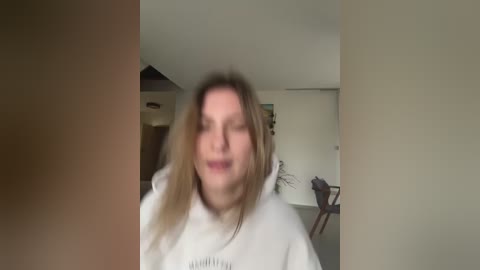 Media: Video of a blurred, middle-aged woman with shoulder-length blonde hair in a white hoodie, taken in a dimly-lit, sparsely furnished living room with beige walls and minimal decor.