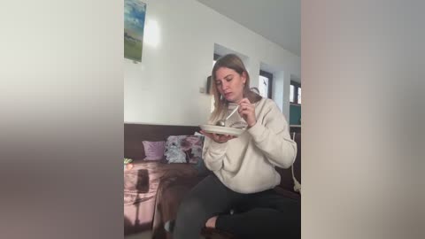 Media: A video of a woman in a cozy living room, wearing a beige sweater and black pants, holding a bowl of soup, with floral pillows and a painting on the wall in the background.