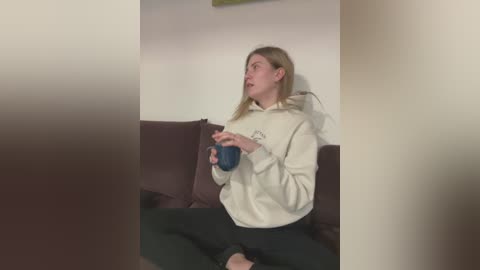 Media: Video of a Caucasian woman with long blonde hair, wearing glasses, a beige hoodie, and black pants, sitting on a brown couch, holding a blue mug, looking upward.