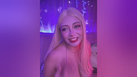 Media: Video of a young, light-skinned, blonde woman with straight hair, wearing red lipstick, smiling, in a room with purple and blue lights. She appears topless, with a hint of her breasts visible.