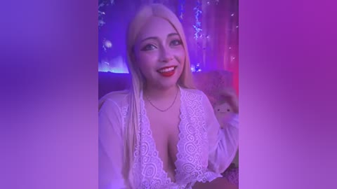 Media: Video of a smiling, fair-skinned woman with long blonde hair, wearing a white lace top, in a dimly lit room with purple and blue lights.