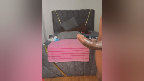 Media: A video of a man's erect penis standing beside a gray, quilted bed with a pink blanket and a dark headboard. The background is a simple, dimly lit room with a wooden floor.