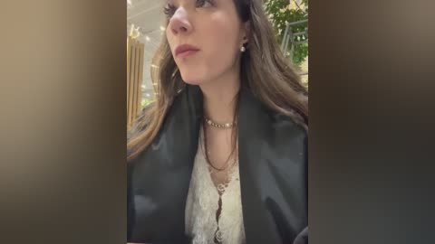 Media: Video of a woman with long brown hair, wearing a black leather jacket over a white lace top, standing indoors near a golden chandelier and green plants, looking thoughtful.