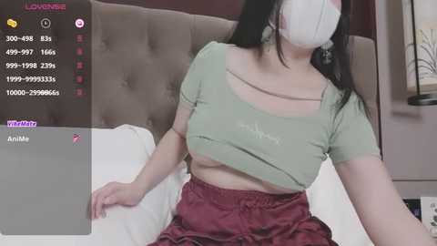 Media: A video of an Asian woman with fair skin and long black hair, wearing a white mask, green top, and red skirt, seated on a bed in a dimly lit bedroom.