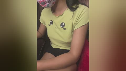 Media: Video of a young woman with medium brown skin, wearing a yellow Pikachu t-shirt and black shorts, sitting on a red cushioned chair. Her face is partially covered by a pink mask with a cartoon character.