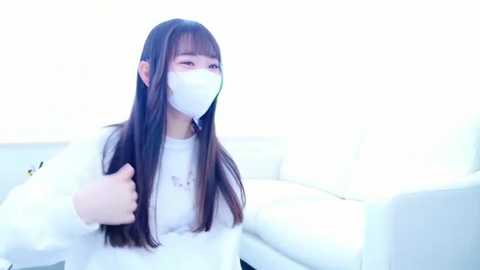 Media: Video of an East Asian woman with long black hair, wearing a white face mask, white jacket, and gloves, sitting in a white room with bright, diffused light.