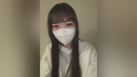 Video of an Asian woman with long, straight black hair, wearing a white surgical mask, light brown top, and beige wall background.