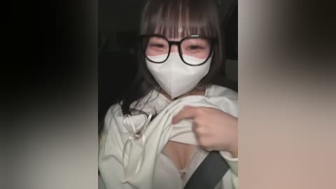 Media: Video of an East Asian woman with straight, black hair and bangs, wearing a white mask, black-framed glasses, and a white hoodie. She is lifting the hoodie, revealing a white bra. The background is dark and blurred.