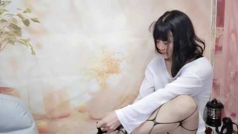 Media: Video of a young woman with long black hair, wearing a white shirt and black fishnet stockings, sitting on a tiled floor, fiddling with her footwear, in a pastel-colored room with floral wallpaper.