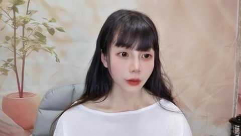 Media: Video of a young East Asian woman with long black hair, pale skin, and light makeup. She wears a white top, standing indoors with a beige wall and potted plant in the background.