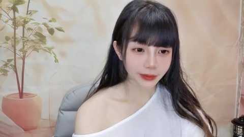 Media: Video of a young East Asian woman with long black hair and fair skin, wearing a white off-shoulder top, seated in a room with a beige wall and a potted plant in the background.