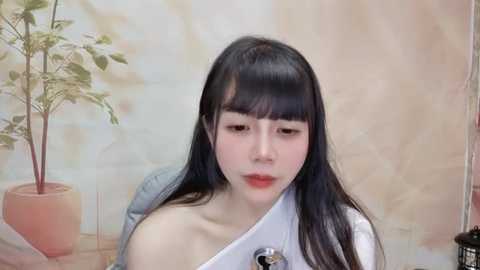 Media: Video of an Asian woman with long black hair, wearing a white off-shoulder top, seated in a room with a beige wall and potted plant.