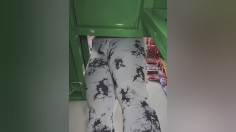 Media: Video of a person's lower body, wearing white leggings with black tie-dye patterns, standing in a brightly lit toy store aisle. The background features green shelving and rows of colorful toy boxes.