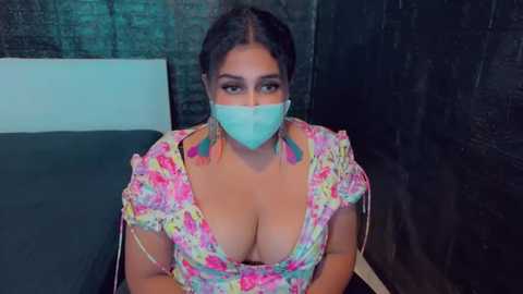 Media: Video of a plus-size woman with light brown skin, wearing a floral-patterned top that reveals cleavage, a face mask, and large, colorful earrings. She sits against a dark brick wall, with a blue bed sheet behind her.