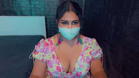 Media: Video of a South Asian woman with medium skin tone and long black hair, wearing a colorful floral dress, blue face mask, and turquoise earrings, seated on a bed in a dark room.