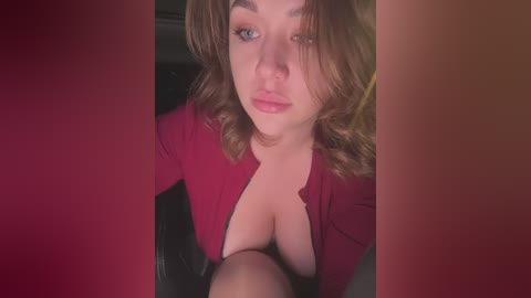 Media: Video of a young woman with wavy brown hair, fair skin, and blue eyes, wearing a plunging red top, revealing ample cleavage. She gazes directly at the camera. Background is blurry, with a red gradient filter.