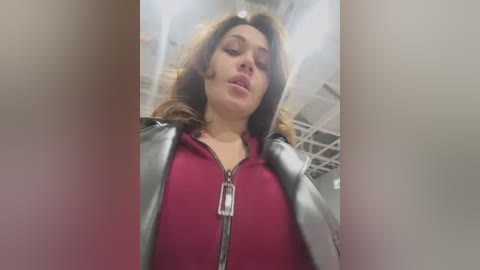 Media: Video of a woman with light skin and wavy brown hair, wearing a red top and black leather jacket, standing in a modern, industrial building with white walls and metal beams.