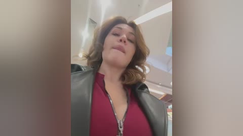 Media: Video of a Caucasian woman with light skin, wavy shoulder-length brown hair, wearing a red top and a black leather jacket, looking confident, in a modern indoor setting with white walls and ceiling lights.