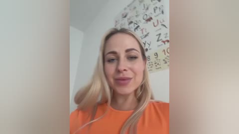 Media: Video of a smiling, fair-skinned woman with long blonde hair, wearing an orange shirt, standing in front of a wall covered with a colorful alphabet chart.