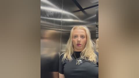 Media: Video of a blonde woman with a surprised expression, wearing a black t-shirt, standing in a shiny, metallic elevator.