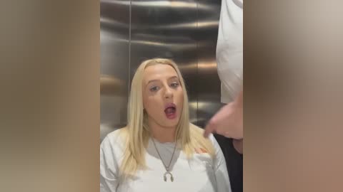 Media: Video of a shocked blonde woman with fair skin, wearing a white t-shirt and necklace, in a metal elevator.