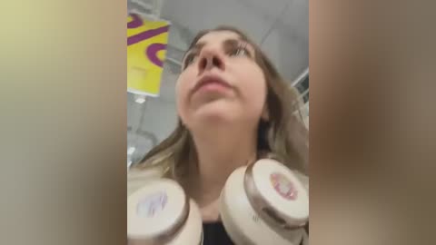 Media: A video of a young woman with light skin and shoulder-length brown hair wearing large white headphones, taken from a low angle, showing her from the waist up. The background is a blurred indoor setting with yellow and purple decorations.