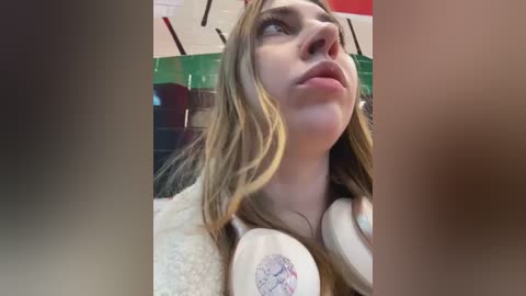 Media: Video of a young woman with long blonde hair, wearing white headphones, looking upward, indoors with green and white walls.