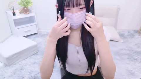 Media: Video of an East Asian woman with long black hair and pale skin, wearing a white mask and a pink top, kneeling on a grey carpet in a minimalistic white room with a white chair.