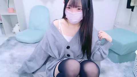 Media: Video of an Asian woman with long black hair, wearing a gray cardigan, white mask, and thigh-high stockings, seated on a fluffy rug in a pastel-toned, minimalist room.