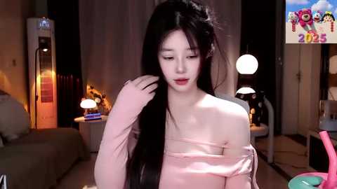 Media: Video of a young East Asian woman with long black hair, wearing a pink off-shoulder dress, touching her hair in a dimly lit bedroom with warm lighting and colorful wall art.