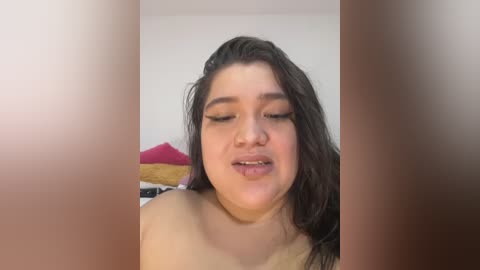 Video of a plus-sized woman with wet, shoulder-length brown hair, wearing no shirt, smiling seductively. Background shows a bed with a pink pillow and a beige blanket.