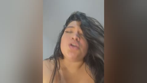 A blurry video of a plus-sized woman with long, dark hair, closed eyes, and a soft expression, possibly in a bedroom setting.