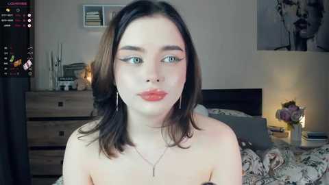 Media: Video of a young, pale-skinned woman with shoulder-length dark hair, wearing red lipstick, sitting topless in a bedroom with floral bedspread, wooden dresser, and framed art on the wall.