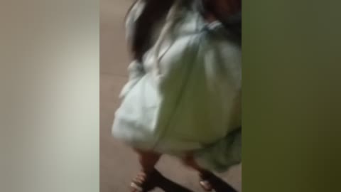 Media: A blurry video of a person in a light-colored dress, possibly green, standing in a dimly lit room with a beige wall. The image is out of focus.