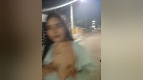 Media: A blurry video of a woman with long black hair, wearing a light blue top, partially exposing her breasts, standing in an urban street at night.