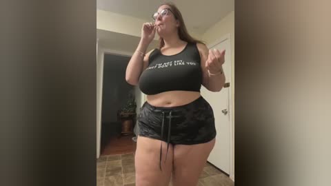 Media: Video of a plus-size woman with glasses and long brown hair, wearing a black crop top and black shorts, in a hallway, striking a pose.