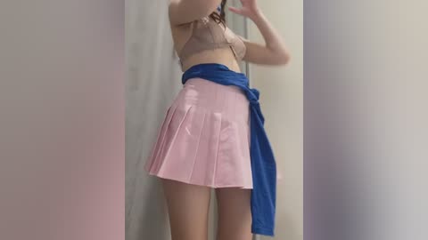 Media: Video of a light-skinned woman with long brown hair, wearing a beige lace bra and a pink pleated mini skirt tied with a blue scarf, standing in a plain, light-colored room.