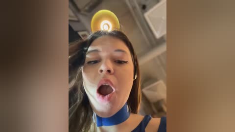 Media: Video of a woman with light brown skin, wearing a blue choker, with her mouth open and a yellow light bulb on her head, in a dimly lit room with metallic structures.