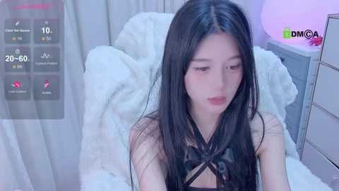 Media: A video of a young East Asian woman with long black hair, fair skin, and a slim physique, wearing a black, strappy top, sitting on a white fur blanket in a softly lit room.