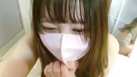 Media: A video of an Asian woman with long brown hair and bangs wearing a white surgical mask, partially obscuring her face, kneeling in a tiled bathroom.