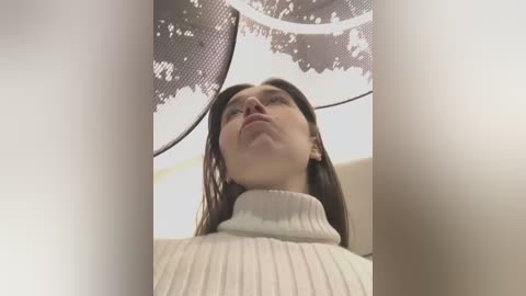 A video showing a young woman with fair skin and straight brown hair, wearing a cream-colored turtleneck sweater, standing under a metal mesh ceiling with a leaf pattern. The image is taken from a low-angle perspective, emphasizing her upward gaze.