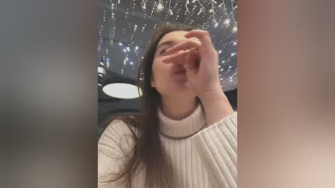 Video of a young woman with long brown hair, wearing a cream-colored knitted sweater, covering her face with her hand in a blurred, candid indoor setting with a festive, twinkling ceiling.