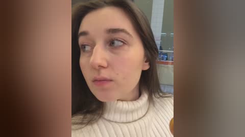 Media: Video of a young woman with fair skin, dark hair, and a neutral expression, wearing a cream-colored turtleneck sweater. Background shows a blurred bathroom with a sink and mirror.