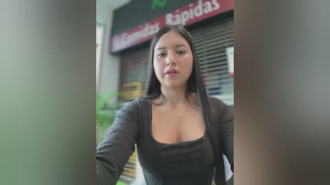 Media: Video of a young, light-skinned Asian woman with long dark hair, wearing a tight, low-cut, black top, standing in front of a closed store with red signage.