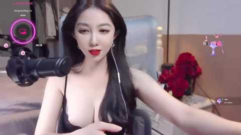 Media: Video of an East Asian woman with long black hair, wearing a black lace bra, singing into a microphone. The room has a modern decor with a bed, a red flower arrangement, and a mirror.