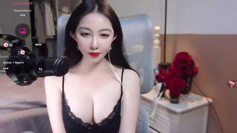 Media: Video of an East Asian woman with pale skin and long black hair, wearing a black lace bra, sitting in a bedroom with a red flower vase and a camera on a tripod.