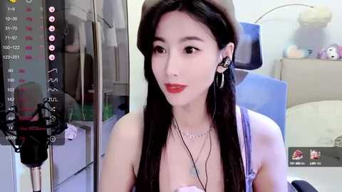 Media: Video of an East Asian woman with fair skin, long black hair, and red lipstick, wearing a beige beret, sitting in a modern room with a blue chair, headphones, and a virtual background.